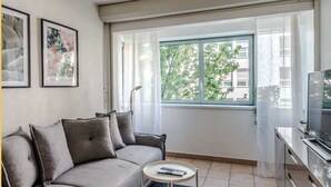 A modern living room with a grey sofa, a small round coffee table, and a TV on a cabinet. There are framed pictures on the wall and a window with a view of trees and buildings.