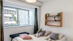 A bedroom with a bed, a bench, a window with curtains, a pendant light, and a wall-mounted shelf with plants.