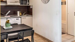 A compact kitchen with a microwave, oven, and sink. A clock is mounted on the wall, and there is a table with chairs.