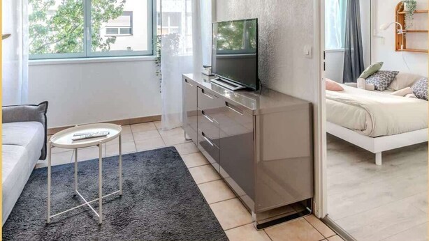 A modern living room with a flat-screen TV on a cabinet, a sofa, a small round table with a remote, and a view of the outside through a large window.
