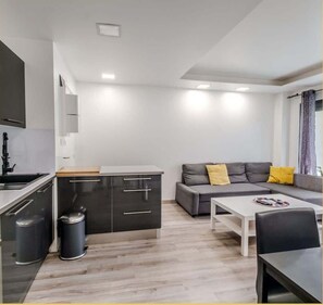 A compact living space with a kitchenette, a sofa with cushions, a dining table, and a trash bin. - Chez Pat' I T2 I Saint-Julien (Saint-Julien-en-Genevois)