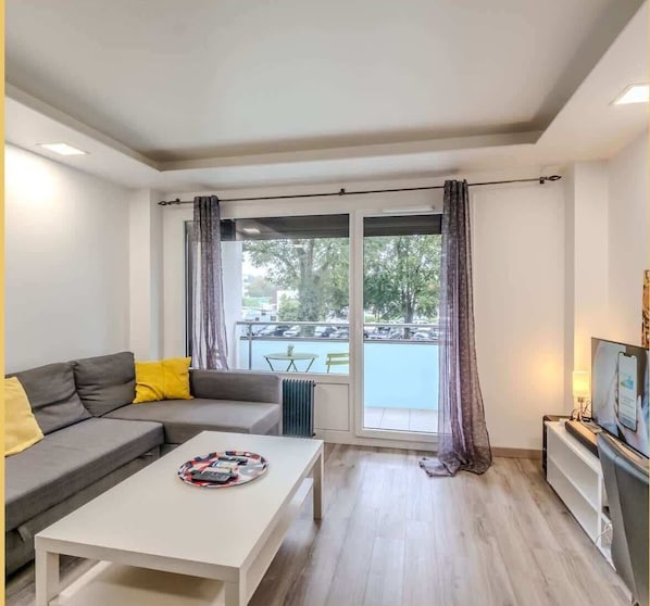 A modern living room with a grey sofa, a white coffee table, and a TV on a stand. A balcony with a table and chairs is visible through the window. - Chez Pat' I T2 I Saint-Julien (Saint-Julien-en-Genevois)