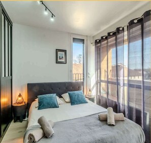 A bedroom with a large bed, two bedside lamps, a window with curtains, and a framed picture on the wall. - Chez Pierre I Studio I Thonon (Thonon-les-Bains)