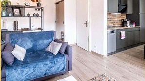 Un salon moderne avec un canapé bleu, un plancher en bois et un coin cuisine doté de meubles gris et d’un réfrigérateur en acier inoxydable.