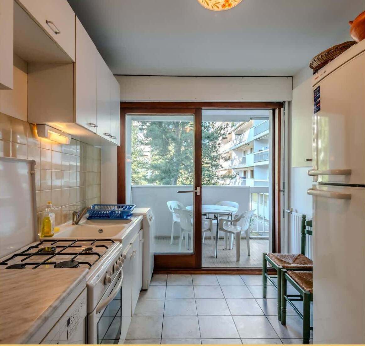 Une cuisine compacte équipée d’une cuisinière à gaz, d’un évier et d’un réfrigérateur. On y trouve également un coin repas avec une table et des chaises, visible à travers une porte vitrée qui donne sur un balcon offrant une vue sur les bâtiments et les arbres.