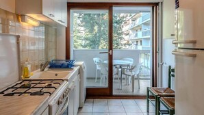 Eine kompakte Küche mit Gasherd, Spüle und Kühlschrank. Durch eine Glastür ist ein Essbereich mit Tisch und Stühlen sowie ein Balkon mit Blick auf Gebäude und Bäume zu sehen.
