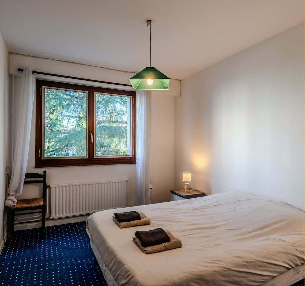 Een slaapkamer met een groot bed, een raam met gordijnen, een radiator, een nachtkastje met een lamp en een stoel.