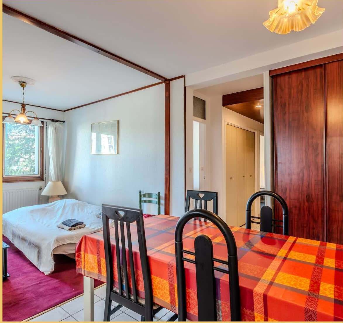 Une pièce comprenant un lit, une table avec des chaises, une fenêtre avec des rideaux et une porte donnant sur une autre pièce.