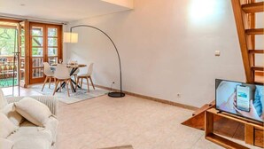 A spacious living area with a high ceiling, a dining table with chairs, a sofa, a floor lamp, and a TV on a wooden stand.
