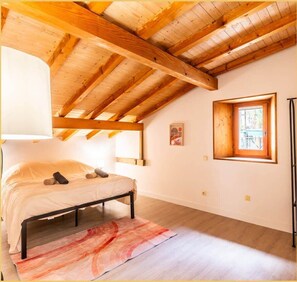 Une chambre avec un plafond en bois, un lit recouvert d’une courtepointe blanche, un tapis rouge et blanc, et une fenêtre donnant sur l’extérieur.