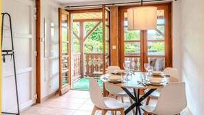 A dining area with a table set for four, surrounded by white chairs, a hanging lamp, and a view of greenery outside.