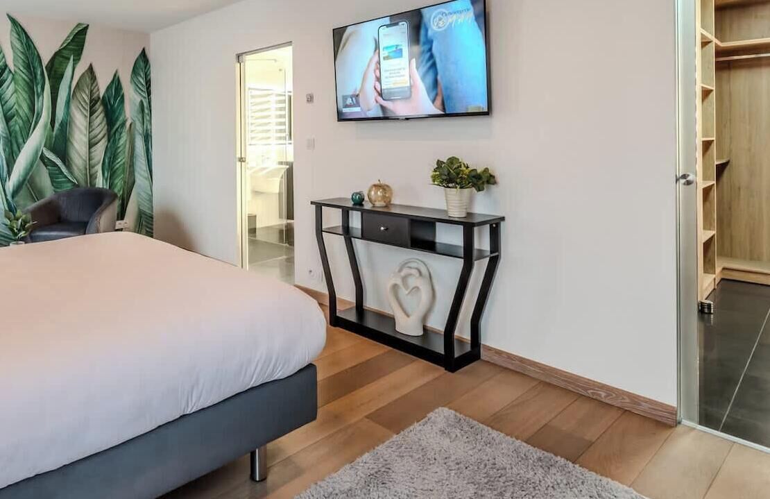 A bedroom with a bed, a TV mounted on the wall, a console table with decorative items, and a large window with a view of the outside.