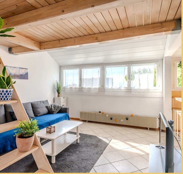 Una sala de estar moderna con un sofá azul, una mesa de centro blanca y un televisor. Hay ventanas, un radiador y un techo de madera.