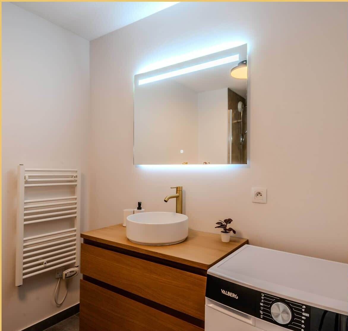 Un baño moderno con un lavabo blanco, un espejo con iluminación integrada, un perchero y un electrodoméstico Valberg.