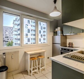 A modern kitchen with a built-in oven, a refrigerator, and a large window with a view of a residential building. There are two stools and a small table in the kitchen area. - Chez Milène I T2 I Ambilly (Ambilly)