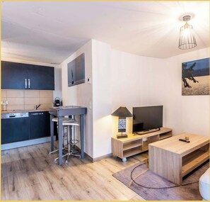 Une cuisine moderne avec des meubles de couleur sombre, un lave-vaisselle et un tabouret de bar. On aperçoit un téléviseur posé sur un meuble en bois, avec une lampe à côté. Un tableau est accroché au mur, et on distingue un luminaire au plafond.