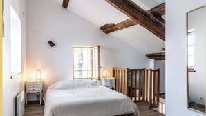 A bedroom with a large bed, wooden beams, a window, a bedside table with a lamp, and a small round table.