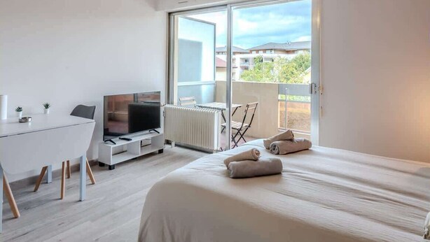 A modern hotel room with a large bed, a desk with a television, and a balcony with a view of buildings.