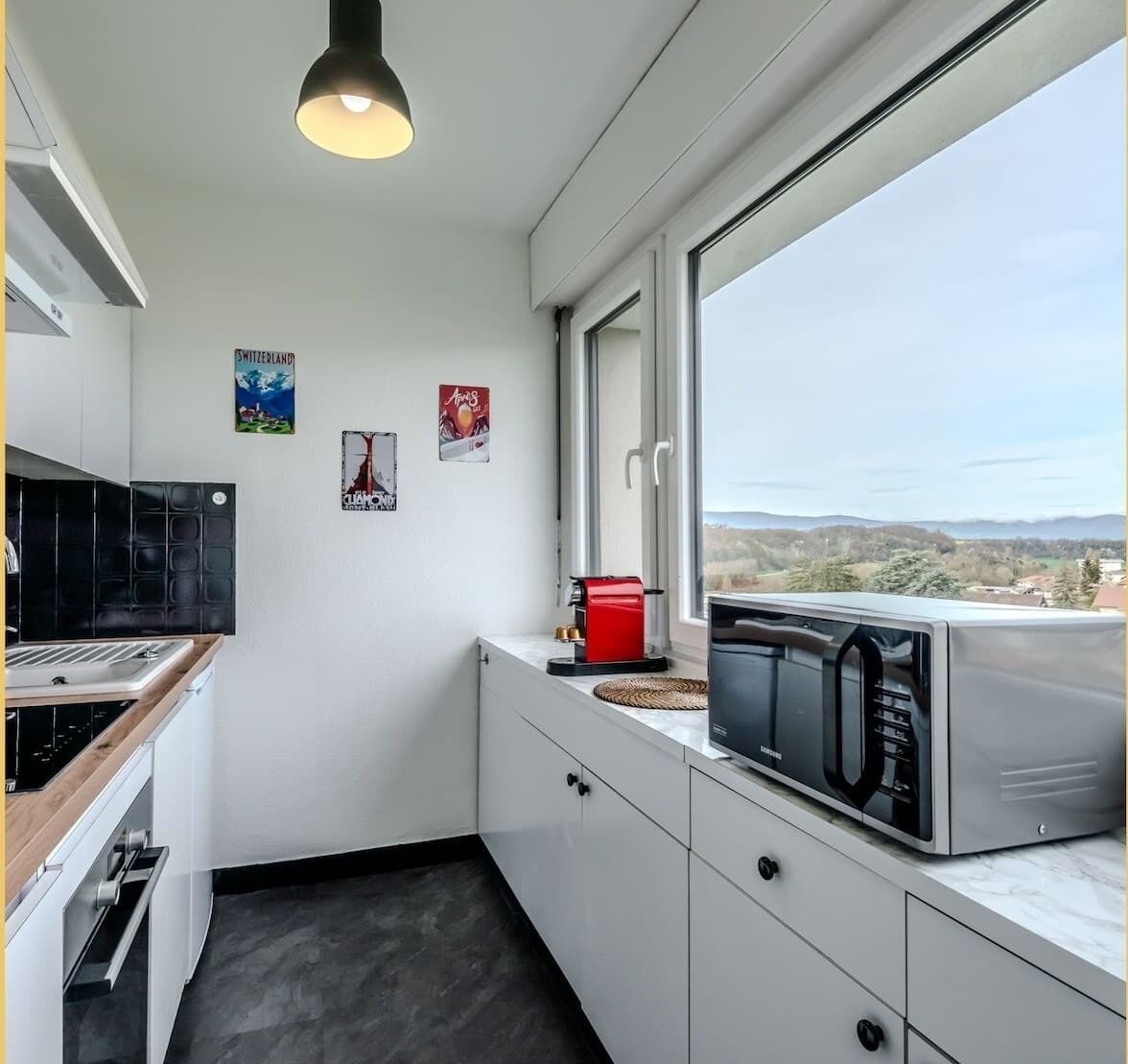 A modern kitchen with white cabinets, a black stove, a red coffee maker, and a window with a view of the countryside.