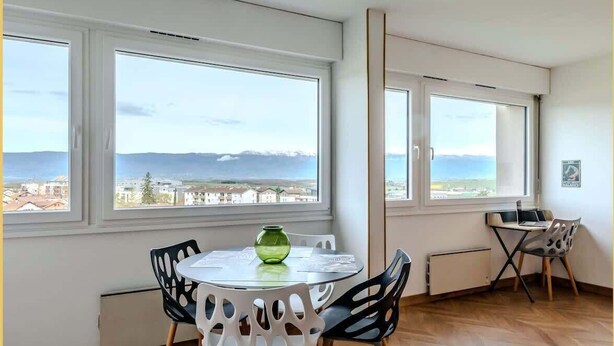A modern room with large windows, a round table with chairs, and a view of the sea and mountains.
