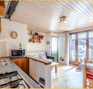 Une cuisine compacte équipée d’un four à micro-ondes, d’une cuisinière et de meubles en bois. On y trouve un petit coin repas avec un banc et une fenêtre donnant sur un balcon.