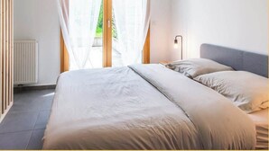A bedroom with a bed, a bedside table, a lamp, and a window with curtains. - Chez Hubert | Proche Genève I Beaumont (Beaumont)