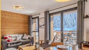 A modern living room with a wooden wall, a grey sofa, a dining table set for four, and a large window offering a snowy outdoor view. - L'évasion I T3 I Les Gets (Les Gets)