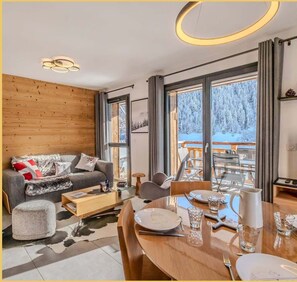 Een moderne woonkamer met een houten wand, een grijze bank, een eettafel gedekt voor vier personen en een groot raam met uitzicht op een besneeerd landschap.