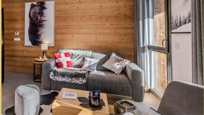 A modern living room with a wooden wall, a grey sofa, a coffee table, and a large window with a view of trees. - L'évasion I T3 I Les Gets (Les Gets)