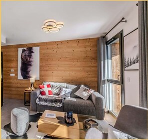 Een moderne woonkamer met een houten wand, een grijze bank, een bijzettafeltje en een groot raam met uitzicht op bomen.