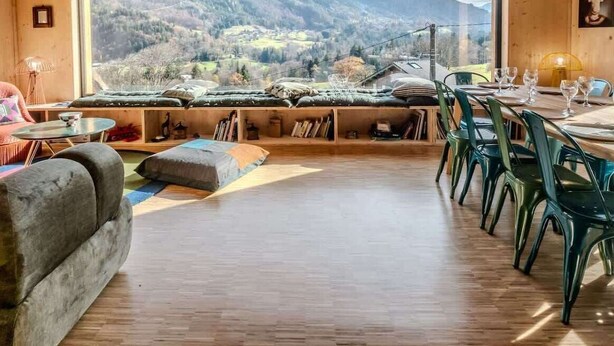 Ein gemütliches Zimmer mit einem großen Fenster und Blick auf die Berge, Holzwände und einer Essecke mit grünen Stühlen.