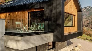 Eine moderne Hütte mit Satteldach, großen Fenstern und einer Beton-Terrasse mit Sitzgelegenheit.