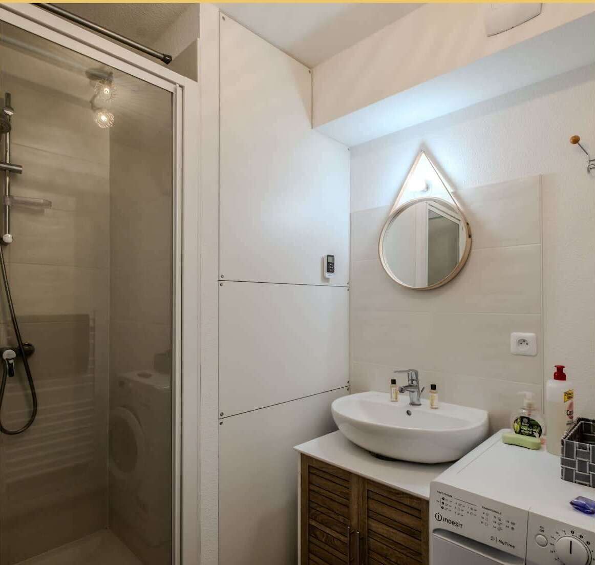 A modern bathroom with a glass-enclosed shower, a round mirror, a white sink, and a built-in washing machine.