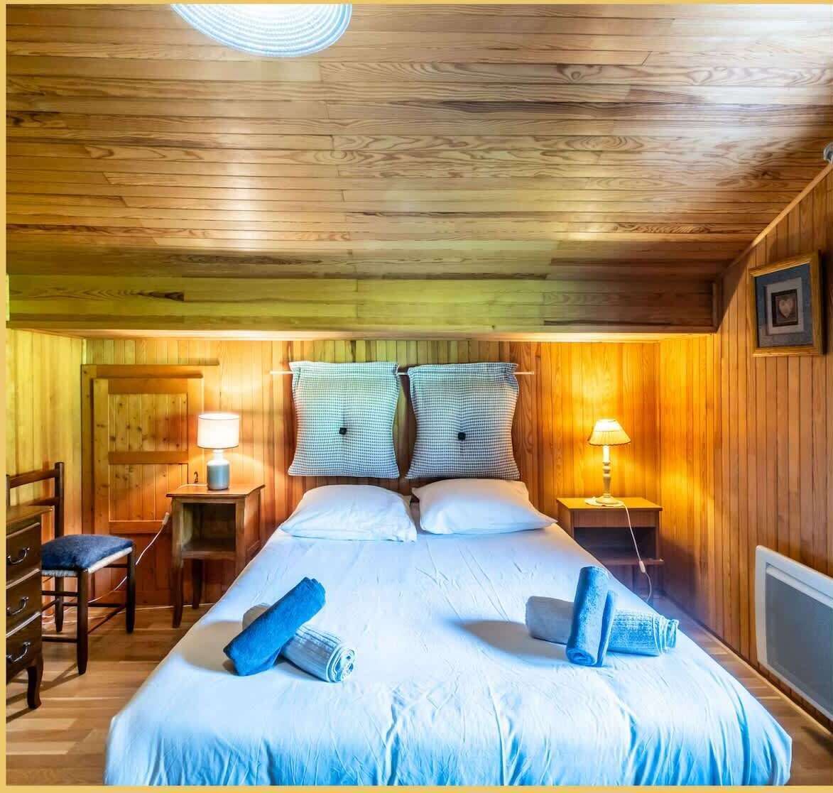 A cozy wooden room with a bed, two bedside tables with lamps, a chair, and a dresser.
