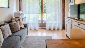 Uma sala de estar acolhedora com teto de madeira, sofá cinza, televisão sobre um móvel e cortinas leves sobre uma porta de vidro.