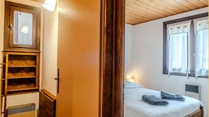 Quarto com teto de madeira, cama com roupa de cama branca, janela com cortinas e guarda-roupa de madeira com espelho.