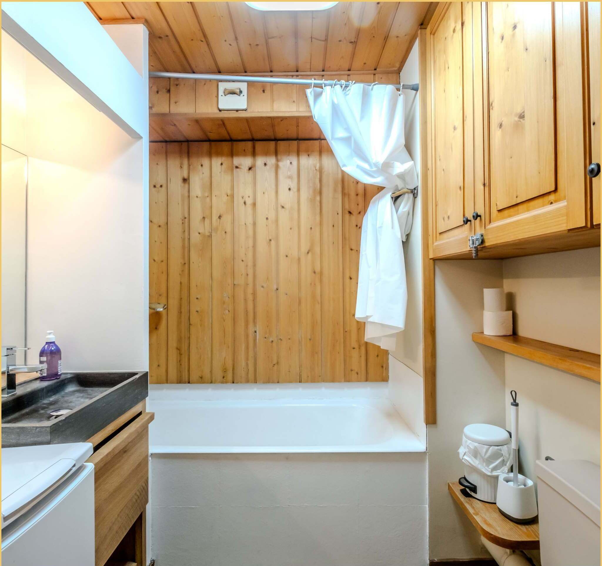 Een kleine, praktische badkamer met houten lambrisering, een bad, toilet en wastafel.