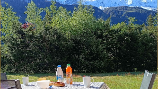 Mesa posta ao ar livre, vista para montanhas, suco de laranja, caneca branca e pão.