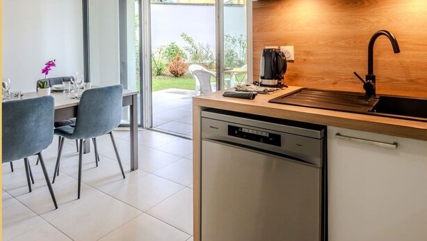 Una cocina moderna con lavavajillas, cafetera y lavabo. Hay una zona de comedor con sillas y mesa, y vista a un jardín a través de un ventanal.