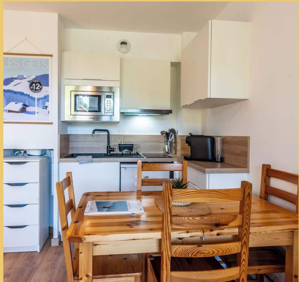 Une cuisine compacte équipée d’un four à micro-ondes, d’une cuisinière et d’un évier, ainsi qu’une table à manger en bois avec des chaises et un petit meuble de rangement.