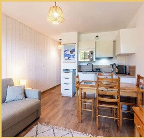 A compact living space with a kitchenette, a dining area with wooden chairs, a sofa, and a hanging light fixture. - Appartement | Sublime | Les Gets (Les Gets)