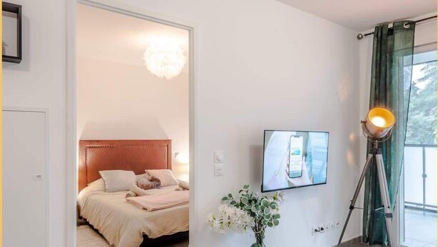 Un dormitorio con cama, televisor montado en la pared, lámpara de pie y un jarrón con flores.