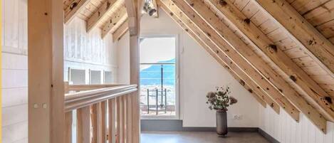 A high-ceilinged room with exposed wooden beams, a balcony with a view, and a potted plant.