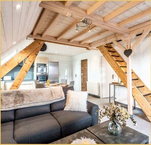 Un espace de vie moderne de style loft, avec des poutres en bois apparentes, un canapé, une table basse et une cuisine en arrière-plan.