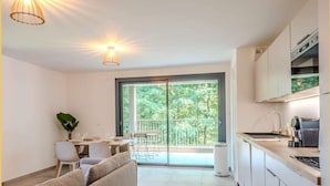 Une cuisine moderne avec un coin repas, un canapé et une porte-fenêtre coulissante donnant sur un balcon avec vue sur les arbres.
