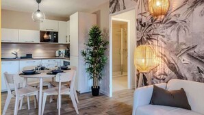 Un salon moderne avec un canapé et un coin repas comprenant une table et des chaises. On y trouve également une plante en pot et un mur orné d’une fresque aux motifs tropicaux.