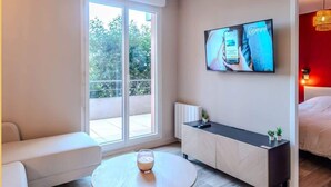 Une chambre d’hôtel moderne avec un canapé, une table ronde en verre, un téléviseur fixé au mur et un balcon offrant une vue sur les arbres.