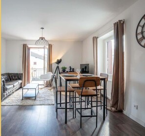 Ein modernes Wohnzimmer mit Sofa, Esstisch und Stühlen. Ein großes Fenster mit Vorhängen, eine Stehlampe und eine Wanduhr sind ebenfalls vorhanden.