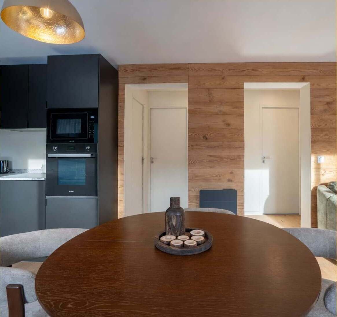 A modern kitchen with dark cabinets, a microwave, and an oven. There's a wooden dining table with a tray of candles.