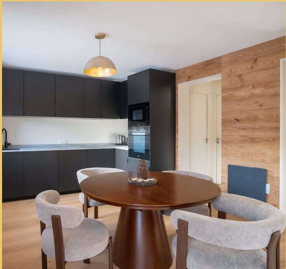 A modern kitchen with dark cabinets, a built-in oven, and a microwave. There is a round wooden dining table with four chairs.